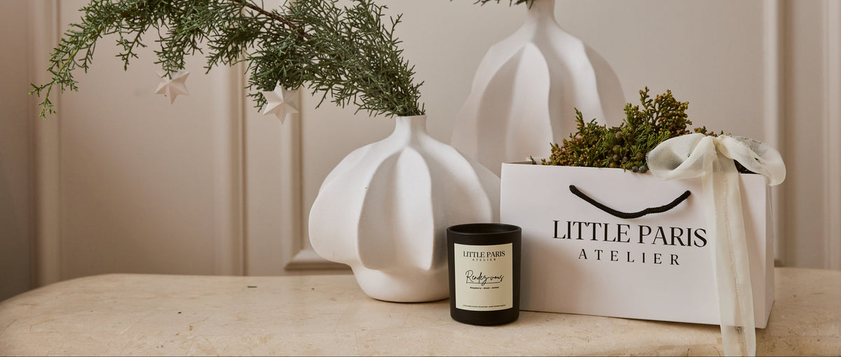 Lumânare Little Paris lângă vase albe din ceramică și o pungă de cadou, în lumina caldă de după-amiază